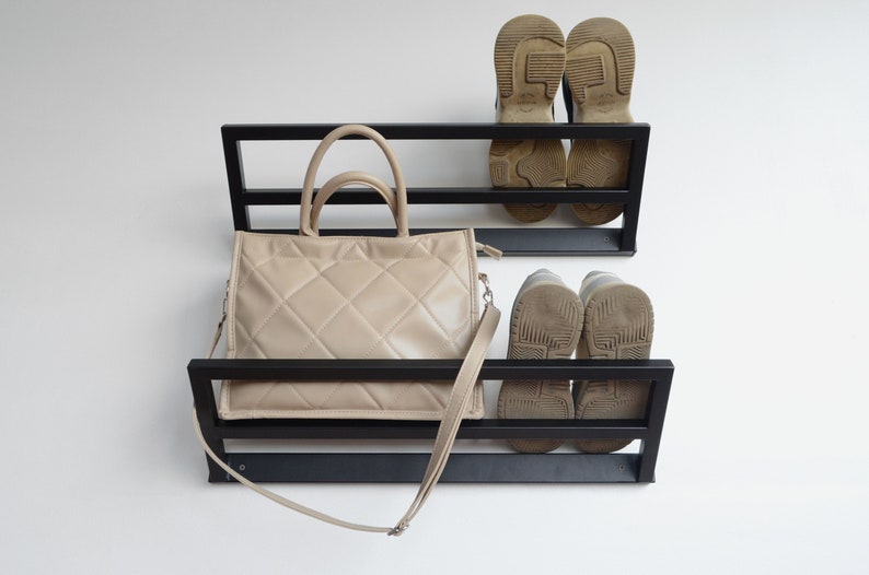 May include: Black metal wall mounted shoe and handbag rack with two shelves. A beige handbag with a long strap is on the top shelf and two pairs of brown shoes are on the bottom shelf.