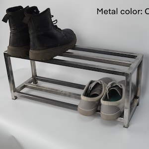 May include: A two-tier metal shoe rack with a clear finish. The top tier has two pairs of black boots, and the bottom tier has two pairs of gray sneakers.