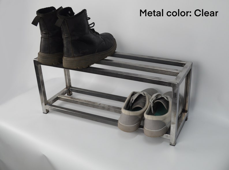 May include: A two-tier metal shoe rack with a clear finish. The top tier has two pairs of black boots, and the bottom tier has two pairs of gray sneakers.