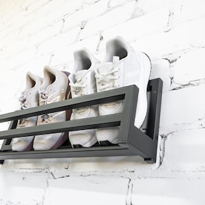 May include: A black metal shoe rack mounted on a white brick wall. The rack has three shelves and is holding four pairs of sneakers.