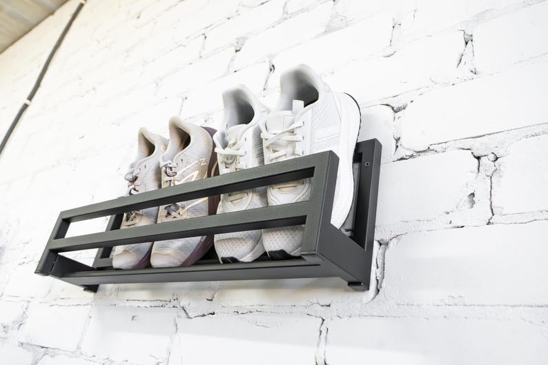 May include: A black metal shoe rack mounted on a white brick wall. The rack has three shelves and is holding four pairs of sneakers.
