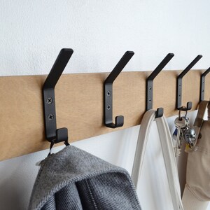 K&ouml;nnte beinhalten: Ein Wand-Kleiderhaken aus Holz mit f&uuml;nf schwarzen Metallhaken. Ein grauer Pullover, eine wei&szlig;e Einkaufstasche, eine beige Baseballkappe und ein Schl&uuml;sselbund h&auml;ngen an den Haken.
