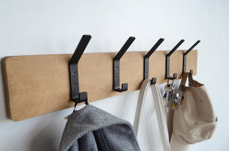 K&ouml;nnte beinhalten: Ein Wand-Kleiderhaken aus Holz mit f&uuml;nf schwarzen Metallhaken. Ein grauer Pullover, eine wei&szlig;e Einkaufstasche, eine beige Baseballkappe und ein Schl&uuml;sselbund h&auml;ngen an den Haken.