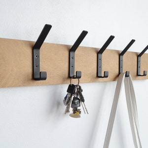 K&ouml;nnte beinhalten: Ein Wand-Kleiderhaken aus Holz mit f&uuml;nf schwarzen Metallhaken. An einem Haken h&auml;ngen Schl&uuml;ssel und an einem anderen eine wei&szlig;e Tasche.