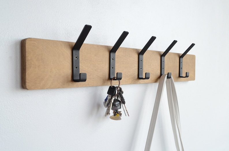 K&ouml;nnte beinhalten: Ein Wand-Kleiderhaken aus Holz mit f&uuml;nf schwarzen Metallhaken. An einem Haken h&auml;ngen Schl&uuml;ssel und an einem anderen eine wei&szlig;e Tasche.