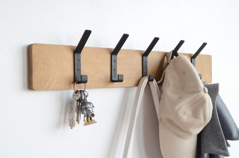 K&ouml;nnte beinhalten: Ein Wand-Kleiderhaken aus Holz mit f&uuml;nf schwarzen Metallhaken. An einem Haken h&auml;ngen Schl&uuml;ssel, an einem anderen Haken ein Hut und eine Tasche und an einem dritten Haken eine Jacke.