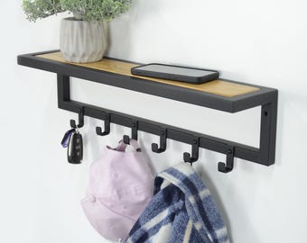 Puede incluir: Estante de pared de metal negro con una parte superior de madera y seis ganchos. Un sombrero rosa, una bufanda de cuadros azules y blancos y llaves cuelgan de los ganchos. Un teléfono móvil está en la estantería.