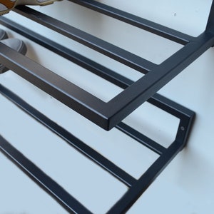 May include: A black metal shoe rack with four shelves mounted on a white wall. The rack is designed to hold shoes and is angled slightly forward.