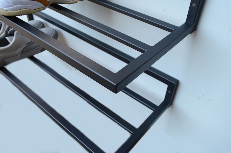 May include: A black metal shoe rack with four shelves mounted on a white wall. The rack is designed to hold shoes and is angled slightly forward.