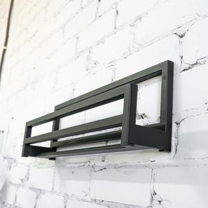 May include: Black metal wall mounted shelf with three tiers. The shelf is mounted on a white brick wall.
