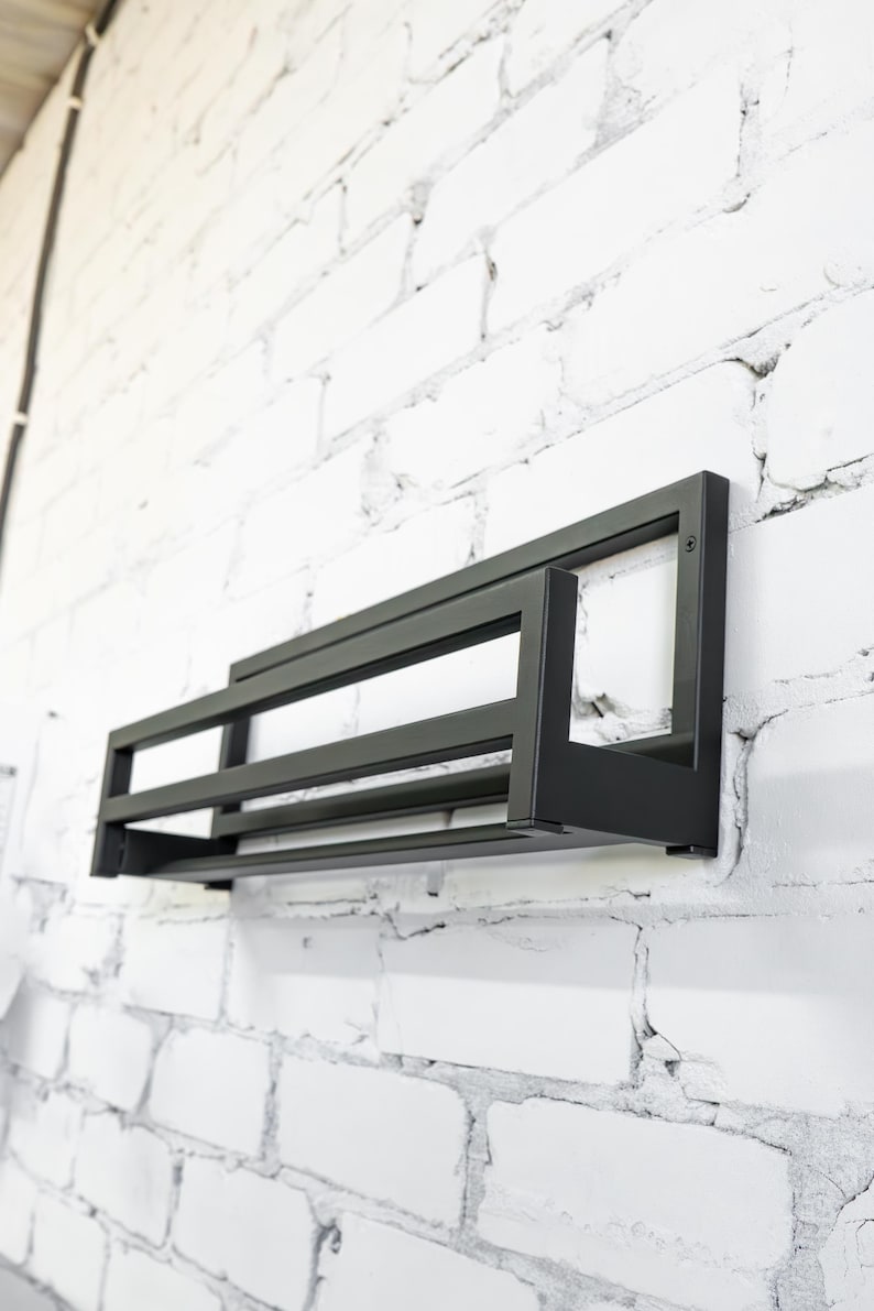 May include: Black metal wall mounted shelf with three tiers. The shelf is mounted on a white brick wall.