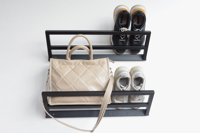 May include: Black metal wall mounted shelf with two tiers. The top tier has two pairs of black sneakers. The bottom tier has a beige quilted handbag with a long strap.