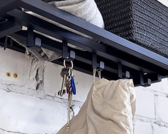 Könnte beinhalten: Schwarzes Metall-Wandregal mit Haken. Eine gefaltete grau-weiß karierte Decke und ein schwarzer Webkorb liegen auf dem Regal. Schlüssel hängen an den Haken, und eine beige Jacke hängt ebenfalls.