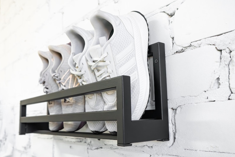 May include: Black metal shoe rack mounted on a white brick wall with three pairs of white sneakers on the shelves.