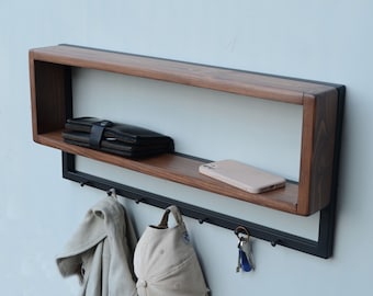 May include: A wall-mounted wooden shelf with a black metal frame and hooks. The shelf has a small open space and is holding a wallet, a phone, and keys. There are also two hooks holding a jacket and a hat.