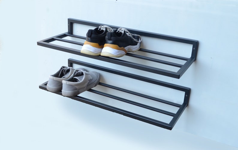 May include: Two-tiered black metal shoe rack mounted on a white wall. The top shelf has two black and white sneakers, and the bottom shelf has two gray sneakers.