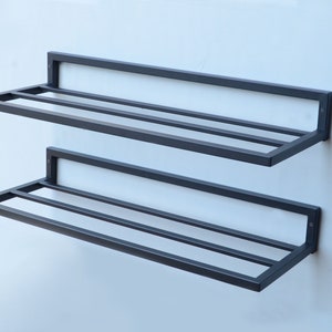 May include: Two black metal shelves mounted on a white wall. The shelves have a simple, rectangular design with horizontal bars for support.