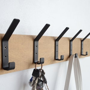 K&ouml;nnte beinhalten: Ein Wand-Kleiderst&auml;nder aus Holz mit f&uuml;nf schwarzen Metallhaken. An einem Haken h&auml;ngen Schl&uuml;ssel und an einem anderen eine wei&szlig;e Tasche.