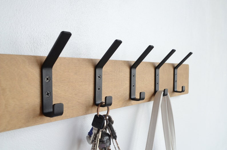 K&ouml;nnte beinhalten: Ein Wand-Kleiderst&auml;nder aus Holz mit f&uuml;nf schwarzen Metallhaken. An einem Haken h&auml;ngen Schl&uuml;ssel und an einem anderen eine wei&szlig;e Tasche.