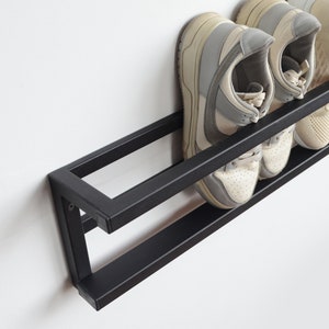 May include: A black metal shoe rack mounted on a white wall, holding several pairs of sneakers. The sneakers are in various colors, including white, gray, and beige. The rack has two levels for shoe storage.