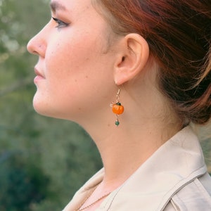 以下が含まれることがあります： 小さなオレンジ色のカボチャのチャームと緑色のビーズが付いたゴールドカラーのイヤリング。カボチャのチャームには緑色の茎があり、ゴールドのフックから吊り下げられています。イヤリングは人の耳に着用されています。