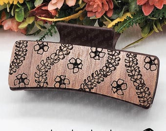 Pode incluir: Um grampo de cabelo de madeira marrom com um design floral. O grampo tem uma forma curva e é decorado com um padrão de flores e folhas. O texto "PUAKENIKENI - LASER TEMPLATE" é visível no topo da imagem.