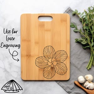 May include: A rectangular bamboo cutting board with a handle, featuring a laser-engraved flower design. The text "Use for Laser Engraving" is visible. The board is displayed on a gray surface with herbs and garlic.