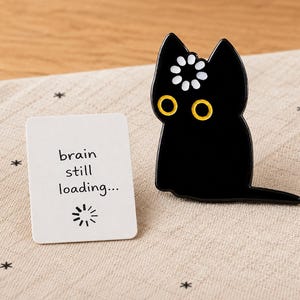 May include: A black enamel cat pin with yellow eyes and a white flower icon on its forehead, positioned next to a rectangular white card featuring the black text "brain still loading..." and a circular loading icon below the words.
