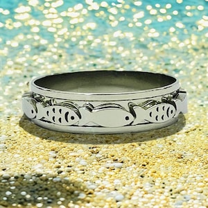May include: A silver ring with a design of fish swimming around the band. The ring is on a sandy surface.