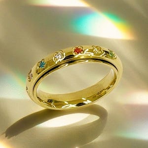 May include: A gold ring with a band of small flowers and colorful gemstones. The ring is on a white surface with a rainbow reflection.