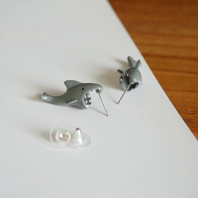 May include: A pair of gray shark-shaped stud earrings featuring an open-mouthed design with visible teeth. The earrings include metal posts and clear plastic earring backs, showcasing a detailed aquatic animal motif in a smooth finish.