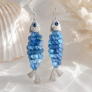 May include: A pair of fish-shaped drop earrings featuring silver-toned metal heads with a hammered texture and blue evil eye beads for eyes. The bodies are composed of layered, circular blue shell discs, and the tails are textured silver-toned metal.