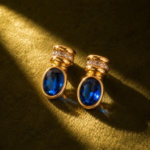 May include: A pair of gold-toned earrings featuring oval-shaped deep blue faceted stones. Each earring has a cylindrical top section embellished with a row of small clear stones, connected to the blue oval base by a ribbed gold-toned band.