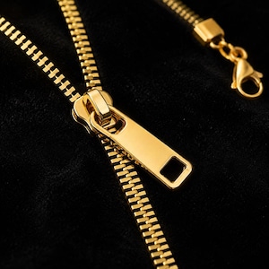 May include: A gold-toned metal zipper necklace featuring a prominent slider pull tab and a lobster claw clasp. The chain is designed to resemble a functional zipper with interlocking teeth, set against a dark black textured surface.