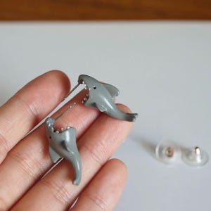May include: A pair of gray shark-shaped stud earrings with open mouths showing white teeth. The earrings feature a smooth finish and include two clear plastic earring backs. The shark design is oriented so the body appears to bite the earlobe.