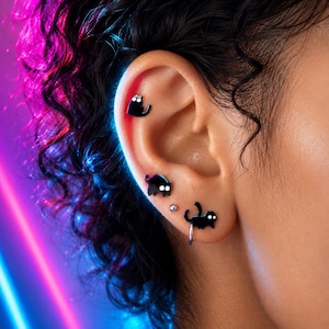 May include: A set of three black cat-shaped earrings with white eyes, including one stud in the upper ear, one stud in the middle, and one dangling cat charm on a small silver hoop earring in the lobe, accompanied by a single small silver ball stud.