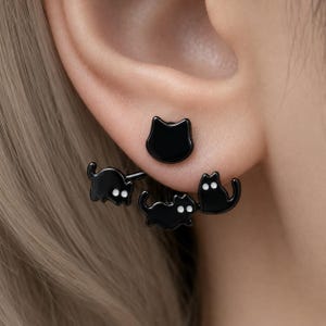 May include: A black cat-themed ear jacket earring featuring a small cat head stud in the front and a larger piece behind the earlobe displaying two additional black cats with white eyes, all crafted with a smooth, glossy finish and metal hardware.