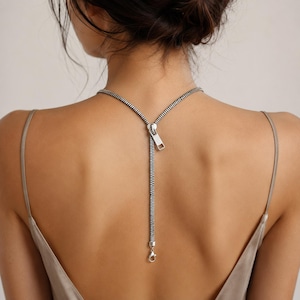 May include: A silver-toned necklace featuring a zipper design that extends down the back. The piece includes a metallic zipper pull and a lobster claw clasp at the bottom, resting against the skin with a Y-shaped configuration.
