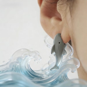 May include: A silver shark earring is worn on an ear. The shark earring is a detailed, miniature sculpture. The background features a clear, blue wave design. The earring is a unique piece of jewelry.