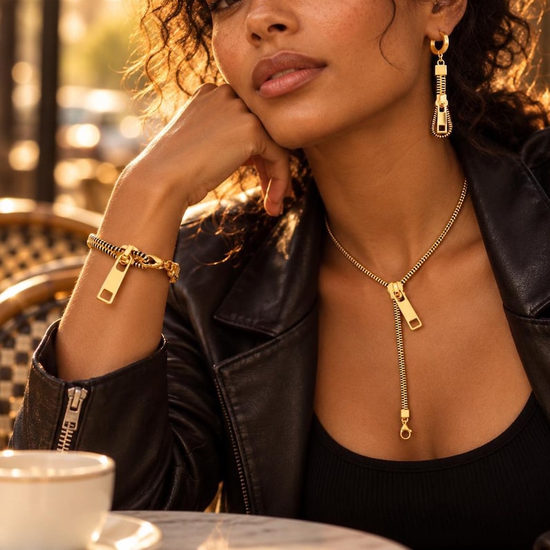 May include: A gold-toned jewelry set consisting of a necklace, a bracelet, and a single earring, all featuring a zipper-inspired design. The necklace has a vertical zipper pull pendant, the bracelet mimics a zipper chain, and the earring is a small hoop with a dangling zipper pull charm.