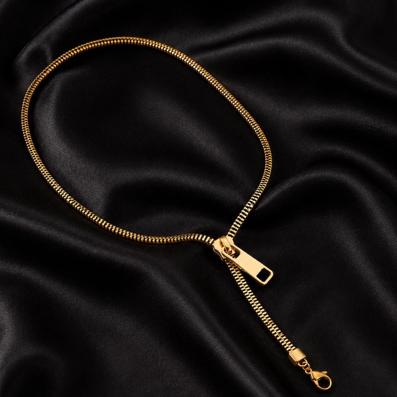 May include: A gold-toned necklace designed to resemble a zipper, featuring a continuous chain of interlocking teeth that form a circular loop, connected to a functional zipper pull tab and a small lobster claw clasp at the end.