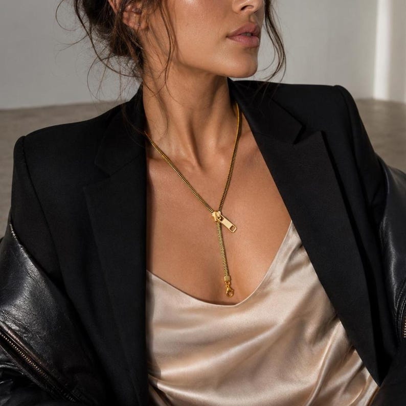 May include: A gold-toned necklace featuring a zipper-inspired design with a functional slider pendant. The chain is composed of interlocking metal teeth that mimic a standard garment zipper, resting against a champagne-colored satin camisole.