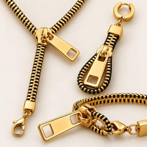 May include: A jewelry set consisting of a necklace, a single earring, and a bracelet, all featuring a design inspired by gold-toned metal zippers with black teeth. Each piece includes a polished gold zipper pull tab and a lobster claw clasp.