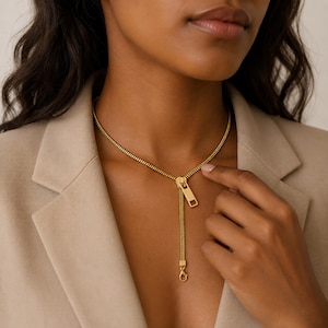 May include: A gold-toned necklace designed to resemble a zipper, featuring a continuous chain that mimics zipper teeth, a central slider component, and a dangling pull tab that terminates in a small lobster claw clasp at the bottom.