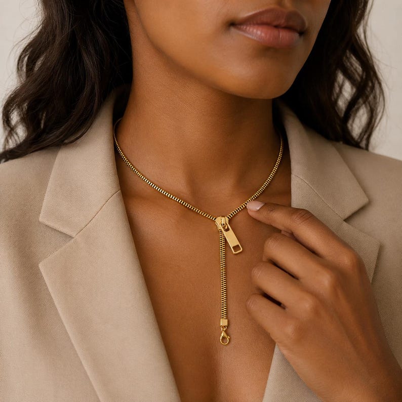May include: A gold-toned necklace designed to resemble a zipper, featuring a continuous chain that mimics zipper teeth, a central slider component, and a dangling pull tab that terminates in a small lobster claw clasp at the bottom.