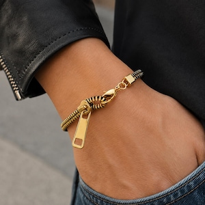 May include: A gold-toned bracelet designed to resemble a zipper, featuring a prominent gold zipper pull tab and a black-and-gold coil chain. The bracelet is secured with a gold lobster claw clasp and includes a small square gold connector piece.