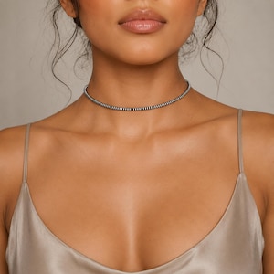 May include: A thin, flexible choker necklace featuring a repeating pattern of small, alternating black and silver-toned metallic beads. The necklace sits closely around the neck, creating a continuous, uniform band of texture and color.