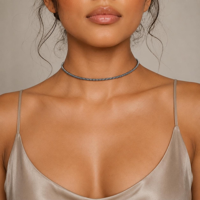 May include: A thin, flexible choker necklace featuring a repeating pattern of small, alternating black and silver-toned metallic beads. The necklace sits closely around the neck, creating a continuous, uniform band of texture and color.