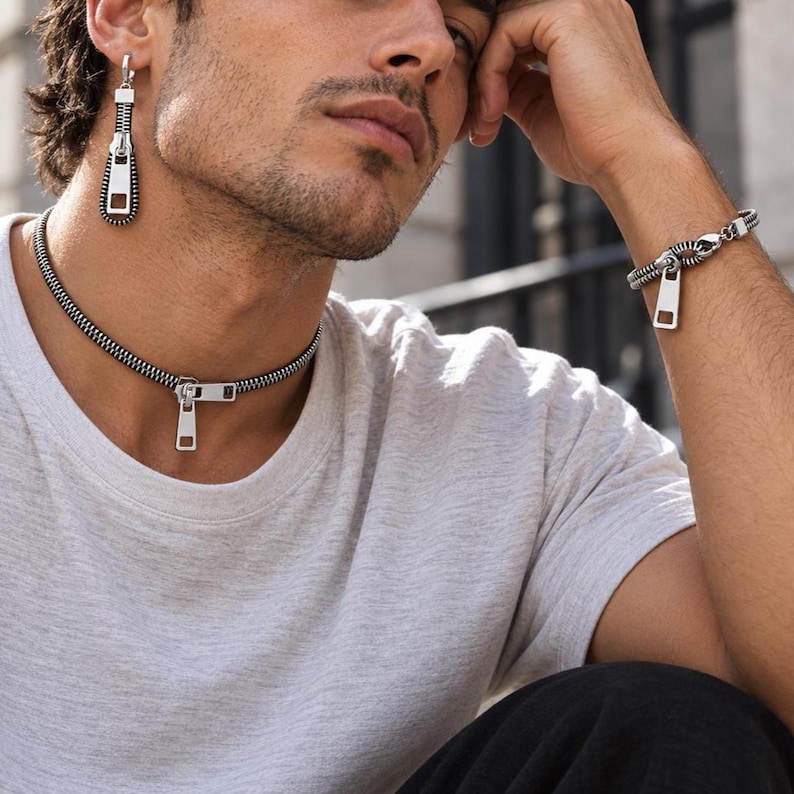 May include: A silver-toned jewelry set consisting of a choker necklace, a single drop earring, and a bracelet. Each piece features a design mimicking a metal zipper with a pull tab, crafted from dark grey and polished silver-colored metal links.