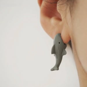 May include: A gray shark-shaped earring worn on an earlobe. The shark is depicted in a vertical orientation with its head pointing upward and its tail pointing downward, featuring a small black eye and visible dorsal and pectoral fins.